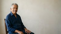Senior man with gray hair and dark skin tone wearing navy blue shirt, seated on chair against neutral background, suitable for healthcare, wellness, and lifestyle editorial use.