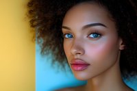 Young woman with medium brown skin tone, curly dark hair, and striking blue eyes wearing pink lipstick against a vibrant yellow and blue gradient background. Suitable for beauty, cosmetics, and lifestyle marketing.