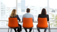 Business professionals seated in orange chairs looking through large office window at urban cityscape, depicting corporate workplace environment and teamwork perspective.