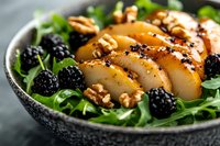 Sliced grilled chicken breast on fresh arugula salad with blackberries, walnuts and honey mustard dressing in dark ceramic bowl on marble surface.