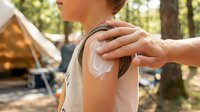 Person applying sunscreen lotion to shoulder outdoors at camping site with tent and forest background for sun protection and skincare.
