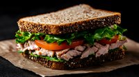 Fresh turkey sandwich with crisp lettuce, juicy tomato slices and dark whole grain bread on rustic brown paper, photographed against black background with dramatic lighting.