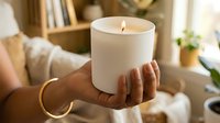 Unlit white candle held in hands of a woman with warm home interior background, aromatherapy and relaxation concept.