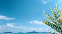 Tropical palm leaf against bright blue sky with white fluffy clouds and distant mountain silhouettes, creating serene summer vacation atmosphere.