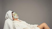 Young woman relaxing in white bathrobe with golden facial mask and towel on head during spa treatment session for skincare and wellness routine.