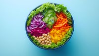 Colorful healthy Buddha bowl with fresh vegetables, chickpeas and greens on blue background. Nutritious vegan meal prep lunch with rainbow salad ingredients.