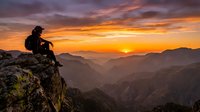 Hiker sitting on rocky mountain peak at golden hour sunrise, overlooking vast valley landscape with layered mountains and misty atmosphere. Perfect for travel, adventure, and outdoor lifestyle content.