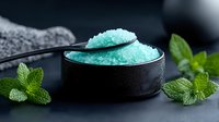 Mint sea salt crystals in black bowl with fresh mint leaves on dark stone surface for spa wellness and aromatherapy treatments.
