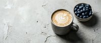 Latte with latte art in gray mug next to bowl of fresh blueberries on textured concrete surface, overhead view for café, food and beverage content.