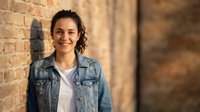 Young Hispanic woman in denim jacket smiling confidently against brick wall background, casual urban portrait for lifestyle and business content.