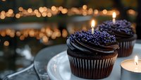 Dark chocolate cupcakes with rich frosting and purple sprinkles topped with lit birthday candles against blurred bokeh lights background on white plate.