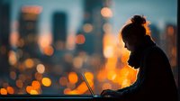 Young professional woman working on laptop computer during golden hour with blurred city lights bokeh background creating atmospheric urban evening scene.