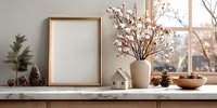 Cozy winter home decor with empty gold frame, cotton branches in vase, pine cones, and miniature house on marble shelf by window. Warm natural light creates inviting atmosphere.