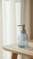 Glass soap dispenser with pump on wooden table near window with natural light filtering through linen curtains in minimalist bathroom interior.