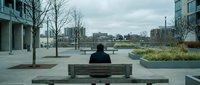 Man in black jacket sitting alone on bench overlooking urban waterfront park with city skyline, modern architecture, and bare trees during overcast day for contemplation themes.