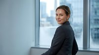 Professional businesswoman in dark suit smiling confidently in modern office with city skyline view through large windows for corporate presentations.