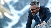 Mature businessman with gray hair and beard in suit climbing rocky mountain peak, demonstrating leadership and determination in challenging environment.