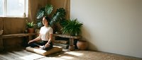 Young Asian woman practicing meditation in lotus position on wooden floor in bright modern room with houseplants creating peaceful zen atmosphere for wellness.
