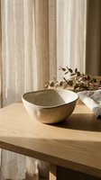Handmade ceramic bowl with speckled glaze on wooden table in warm natural light with eucalyptus branch and linen curtains creating cozy home atmosphere.