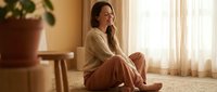 Young woman sitting peacefully on floor by window in warm sunlight, wearing cozy sweater and pants, relaxing at home in comfortable living room setting.