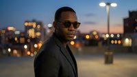 African American male professional wearing sunglasses and dark blazer standing in urban parking area at dusk with city skyline and street lights blurred in background for business and lifestyle use.