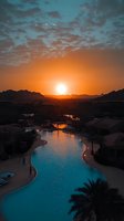 Dramatic sunset over luxury resort swimming pool with mountain silhouettes, glowing orange sky and scattered clouds reflecting in turquoise water, palm trees line poolside.