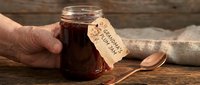 Homemade jam in glass jar with handwritten label held by person's hand, rustic wooden surface with spoon and linen cloth, artisanal preserve for cooking and baking.
