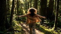 Young woman with curly hair running freely through sunlit forest path with arms outstretched, embracing nature and freedom in lush green woodland setting.