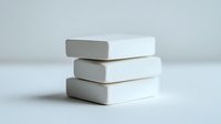 Clean white soap bars stacked in minimalist composition against light background, creating simple geometric shapes and soft shadows. Suitable for wellness concepts.