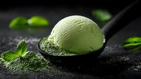 Green matcha ice cream scoop on black spoon garnished with fresh mint leaves against dark background with water drops, close up view for food photography.