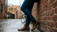 Young man in jeans and brown leather boots leaning against red brick wall on cobblestone street. Urban lifestyle and casual fashion concept for advertising.