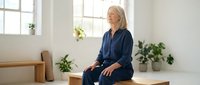 Senior Caucasian woman in navy blue pajamas sitting peacefully on wooden bench in bright minimalist bedroom with plants, morning light streaming through windows.