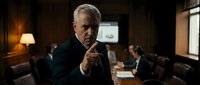 Senior Caucasian businessman in dark suit pointing finger directly at camera in corporate boardroom meeting with colleagues in background.
