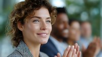 Young professional woman with curly hair and blue eyes smiling warmly during business meeting presentation, with diverse colleagues applauding in background, workplace success moment.