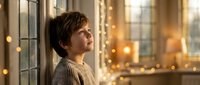 Young boy in cozy sweater gazing thoughtfully out window with warm bokeh lights creating magical atmosphere for holiday season and winter comfort themes.