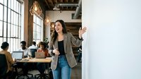 Young Hispanic businesswoman presenting ideas on whiteboard in modern coworking office space with diverse team working in background during meeting.