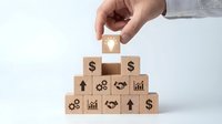 Hand placing wooden cube with lightbulb icon on pyramid of blocks featuring financial symbols including dollar signs, charts, and growth arrows.