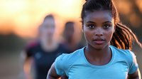 Young African American female athlete in blue shirt running outdoors during golden sunset with training partners in background, focused expression during workout session.