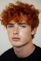 Close-up portrait of young redhead male model with freckles and curly ginger hair wearing black t-shirt against light background, natural daylight.