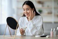 Happy Asian woman applying skincare cream while looking in mirror during morning beauty routine at home vanity table.