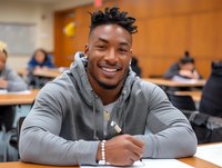 Young African American male student smiling while studying in college classroom, wearing grey hoodie and taking notes, representing education and academic success.