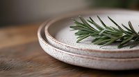 Fresh rosemary sprig on rustic ceramic plates with textured finish, arranged on wooden surface in minimalist composition. Natural light creates soft shadows.