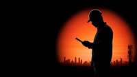 Construction worker silhouette using smartphone against orange sunset with city skyline and communication towers for industrial technology concepts.