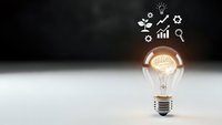 Glowing light bulb with brain inside and business icons floating above on dark background representing innovation and creative thinking concepts.