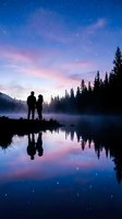 Silhouetted couple standing by misty lake at twilight, gazing at starry sky with pink and purple clouds, evergreen forest reflected in calm water below.