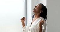 Young African American woman with braided hair wearing a white button-up shirt, smiling with head tilted back and fists clenched in a gesture of triumph, success, and celebration in a bright, minimalist interior space.