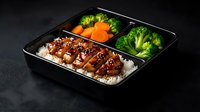 Japanese bento box with teriyaki chicken over rice, steamed broccoli and carrots in compartments on dark background for meal prep and healthy eating.