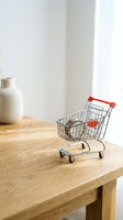 Miniature shopping cart with red handles on wooden table next to white vase, representing retail, commerce, and online shopping concepts for business and e-commerce design.