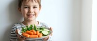 Happy young boy holding plate of fresh vegetables including carrots, cucumbers and greens, promoting healthy eating habits for children and family nutrition.