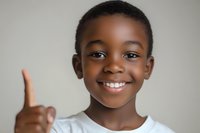 Young African American boy with bright smile showing thumbs up gesture against neutral background, wearing white t-shirt. Positive expression conveys happiness and confidence.